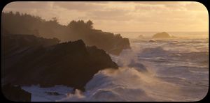 Waves crashing against dark rocky cliffs at golden hour with misty ocean spray